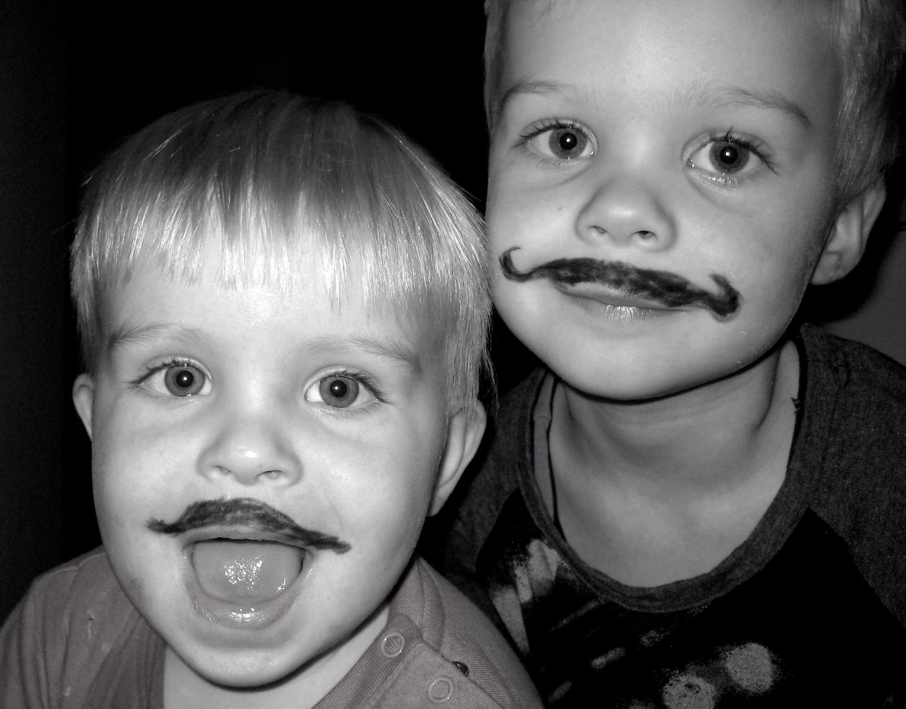 Two young children with drawn-on mustaches smile at the camera, one with an open mouth and the other with a grin.