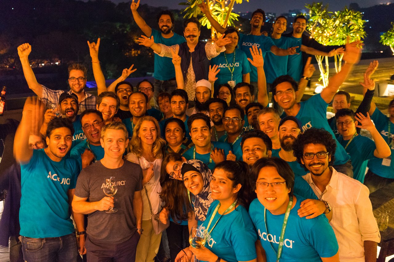 A large group of people in matching blue Acquia shirts gathers outdoors at night, smiling and cheering together.