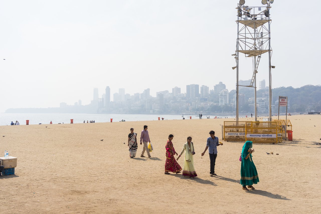 People walk on a sandy beach, some in traditional Indian clothing, with a city skyline visible in the background.