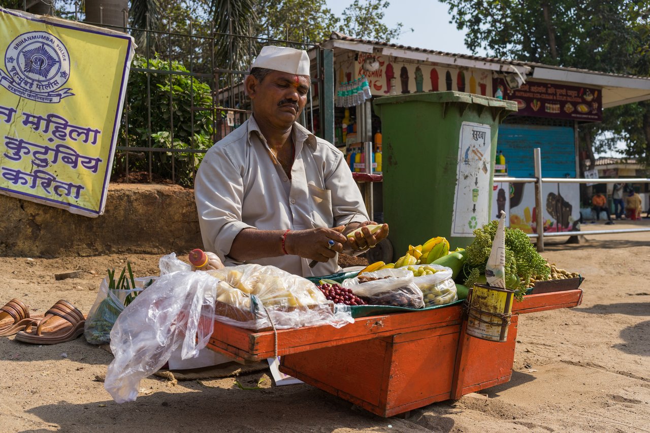 A street vendor in a white cap peels fruit at his small cart filled with fresh produce and snacks.