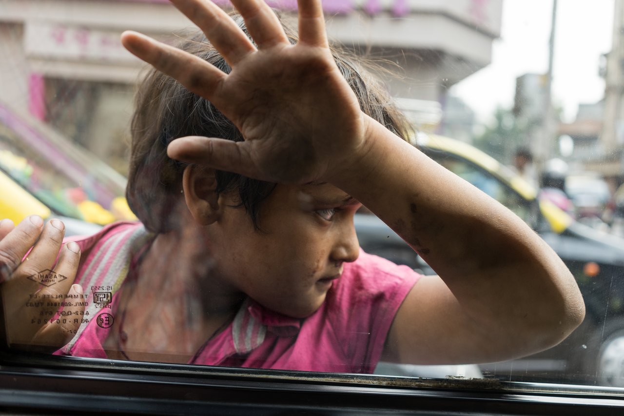 A child in a pink shirt presses against a car window begging for money.