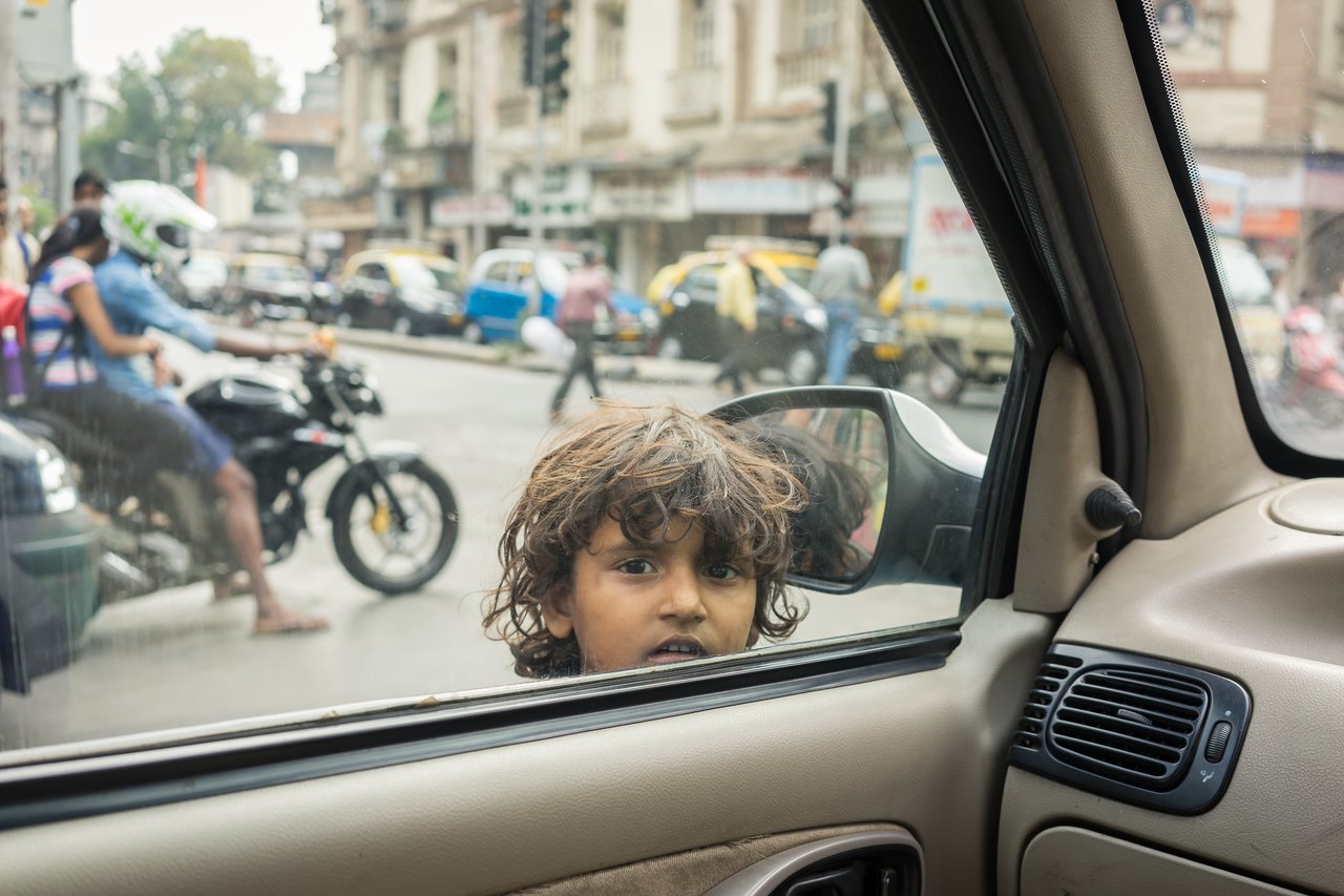 A young child with messy hair looks into a car window begging for money.