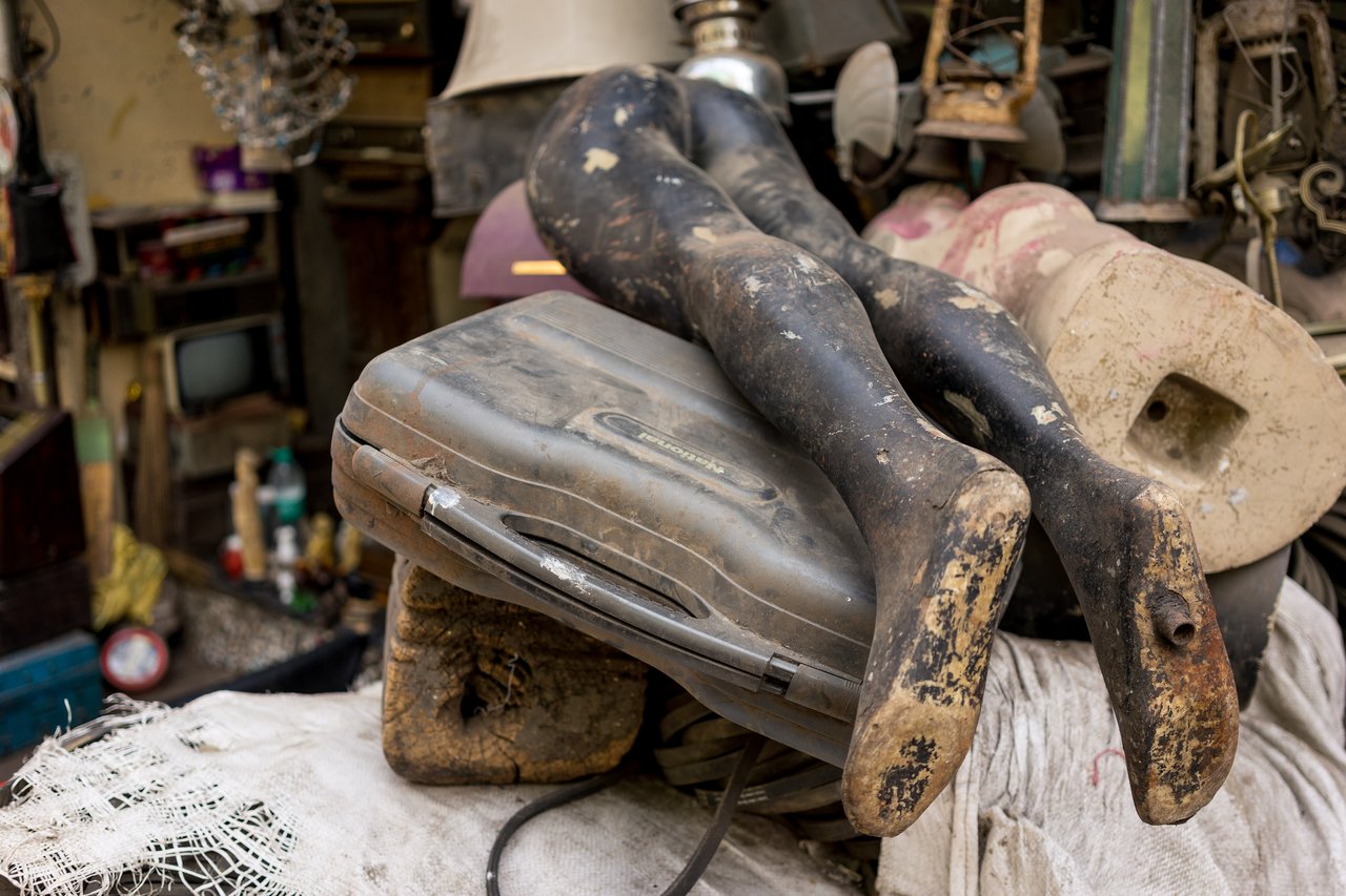 A pair of worn mannequin legs rest on a dusty, old suitcase in a cluttered space.