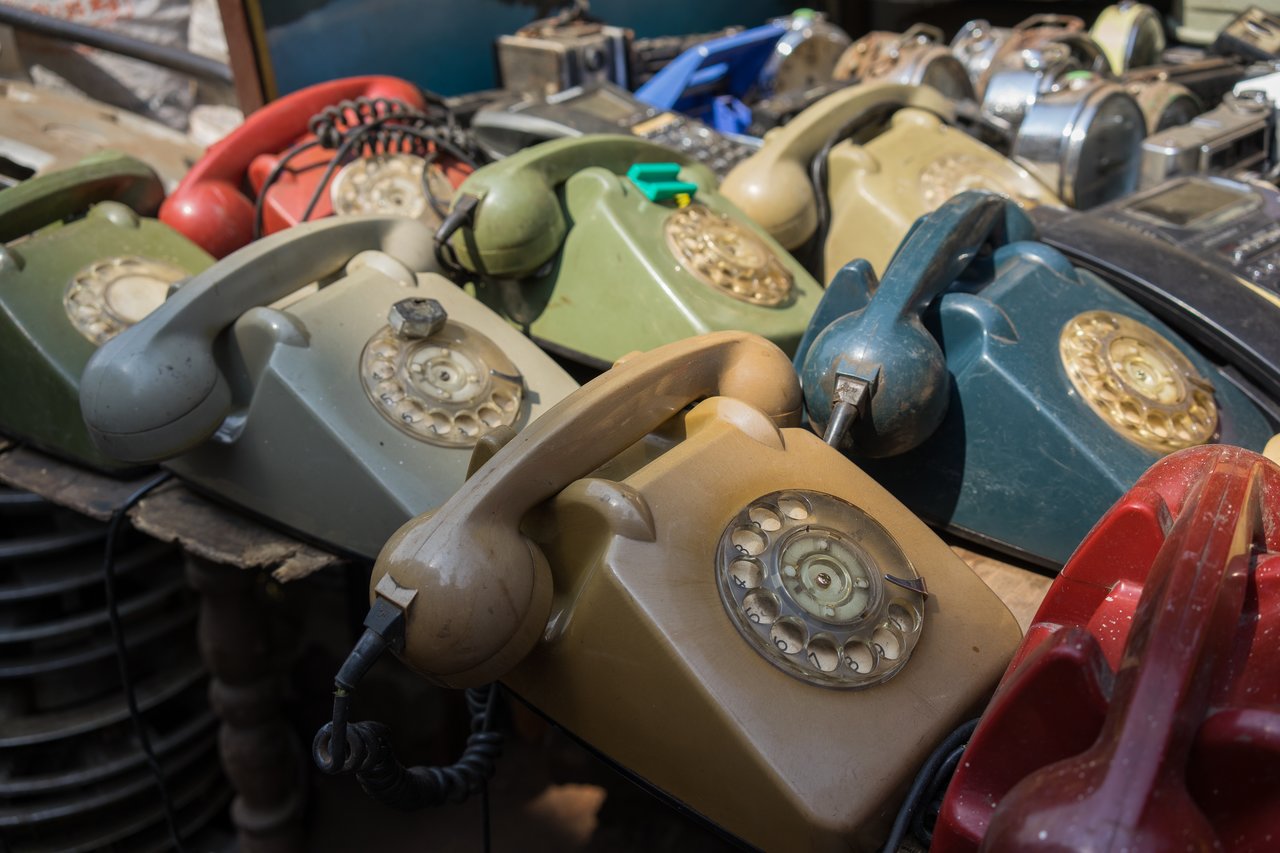 A pile of old rotary telephones in various colors, stacked together on a wooden surface.