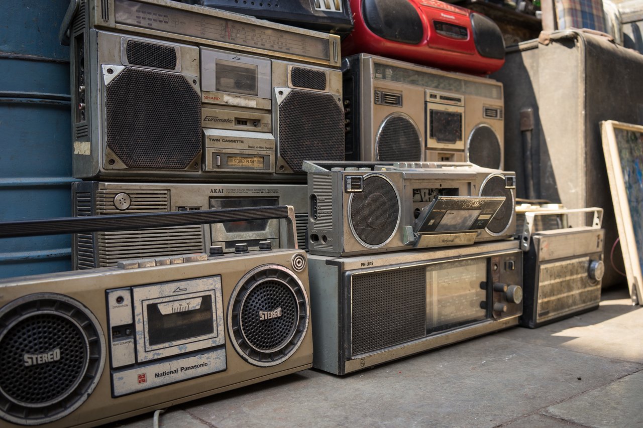 A collection of old, dusty boomboxes and cassette players stacked on the ground, some with open tape decks.