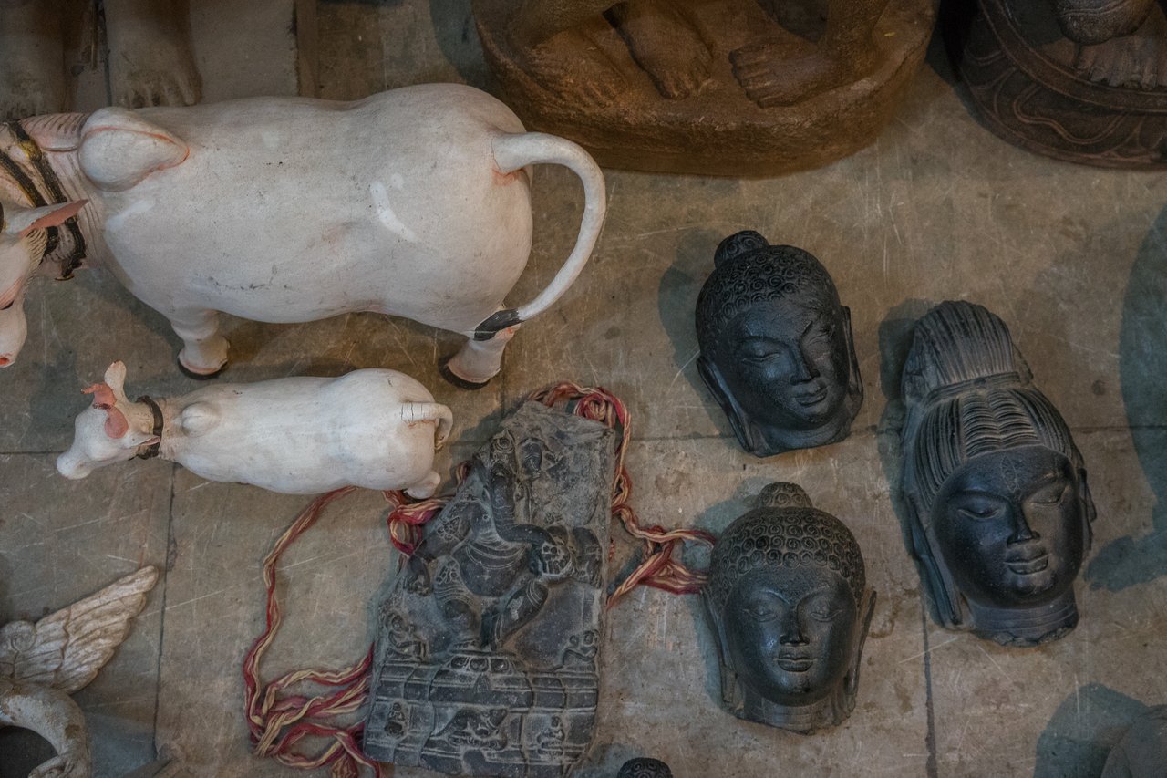 Various stone and ceramic sculptures, including Buddha heads and cow figurines, are displayed on a rough surface.