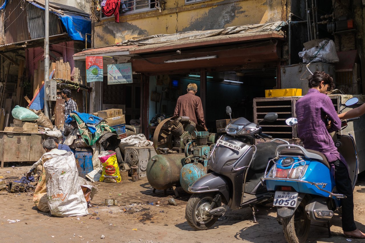 People work outside a repair shop with scattered tools and machinery, while two men sit on parked scooters.