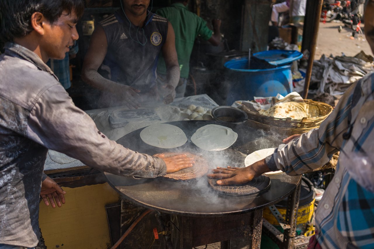 Two people press flatbreads onto a hot griddle with bare hands while another person prepares dough in the background.