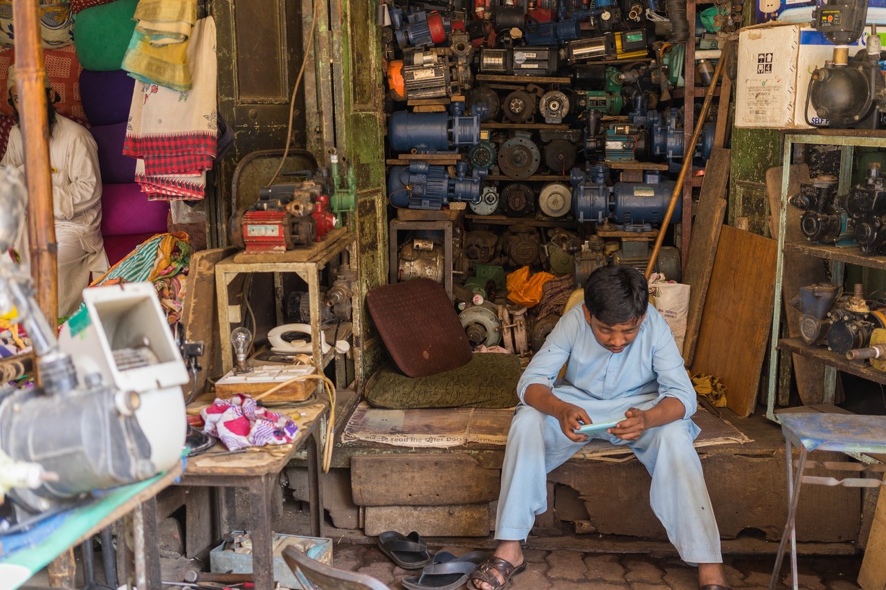 A man in a light blue outfit sits on a bench in a workshop, looking at his phone.