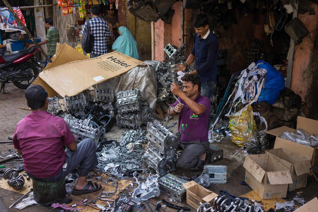 Two men repair and assemble engine parts in an outdoor workshop, surrounded by tools, metal scraps, and cardboard boxes.