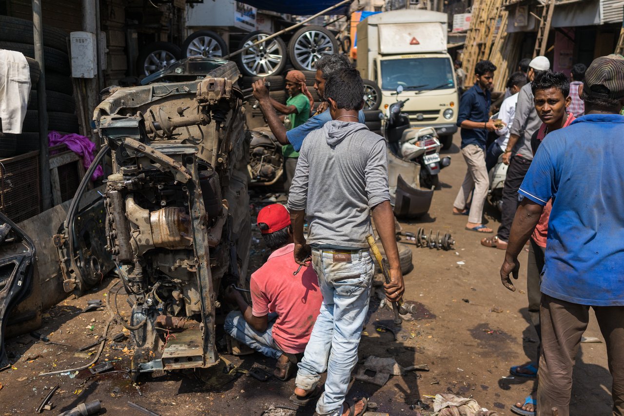 Mechanics work on a dismantled car engine in a busy outdoor repair area, using tools to remove parts.