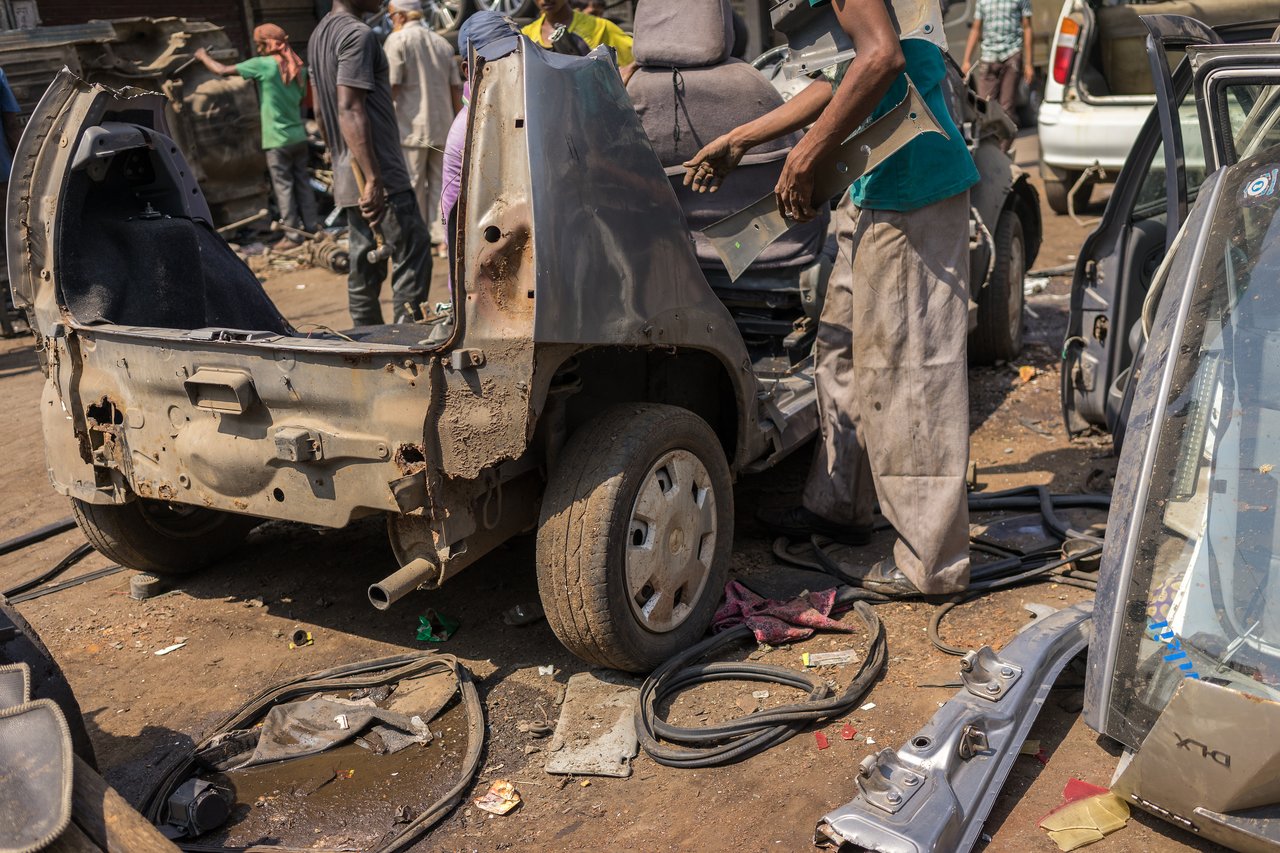 Workers dismantle a damaged car, removing parts and components in an outdoor scrapyard.