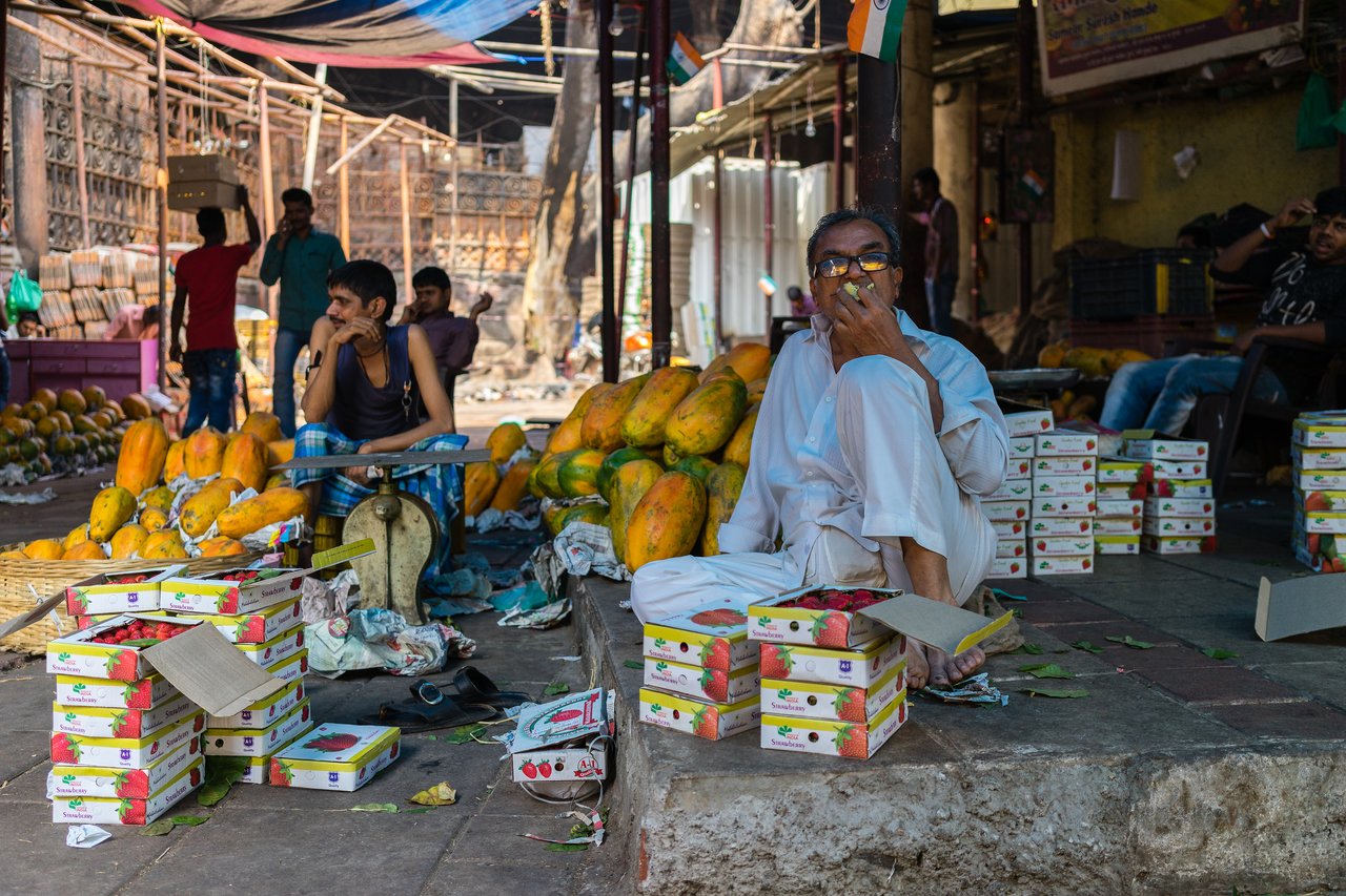A man in white clothing sits on a ledge, eating fruit, surrounded by boxes and papayas at a market.
