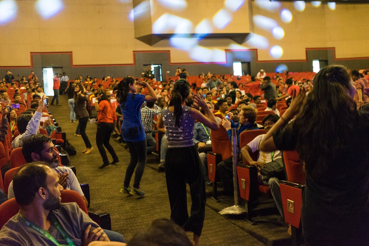 People are dancing in the aisles of a crowded auditorium while others watch, clap, and take photos.