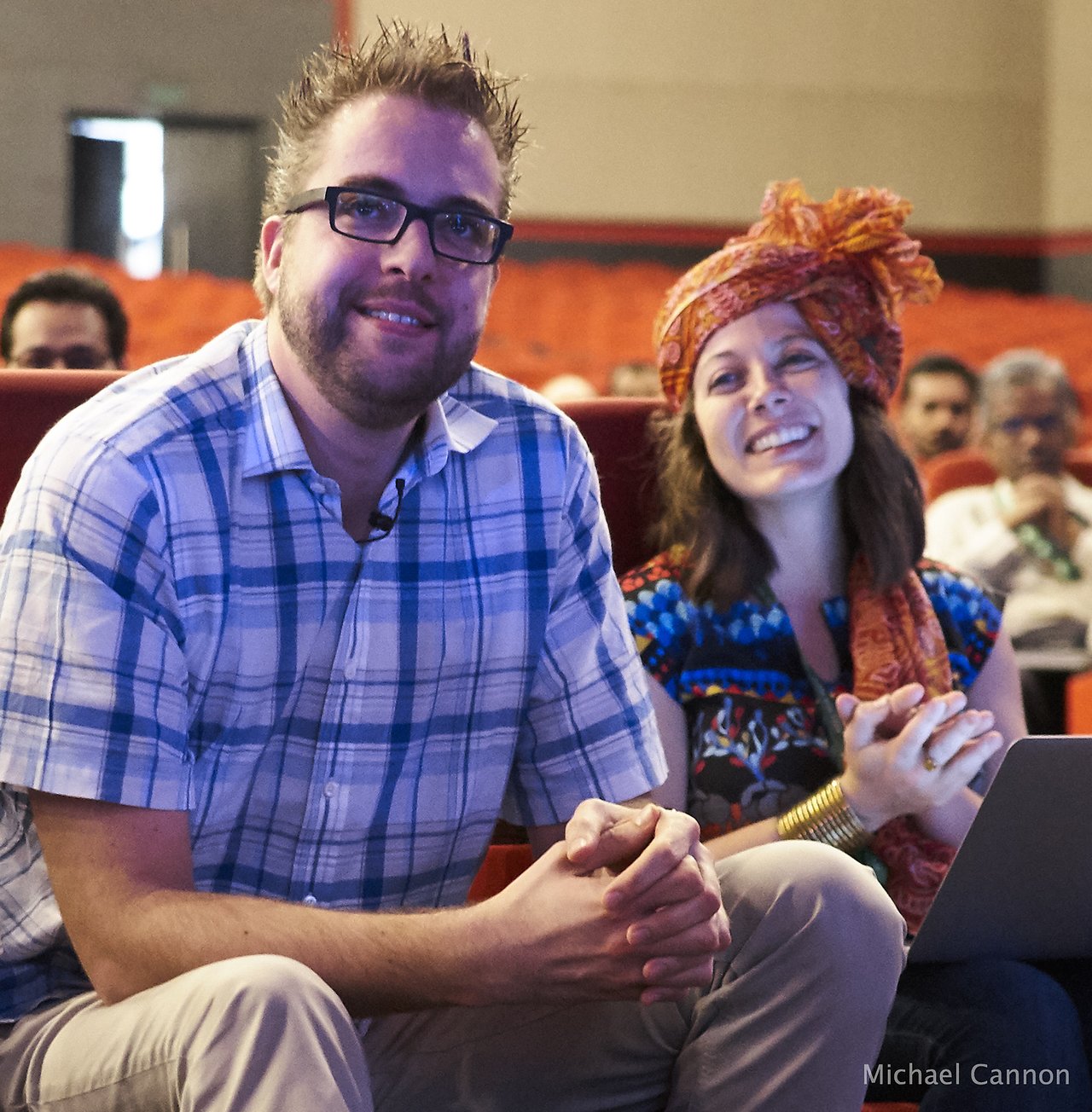 A man in a plaid shirt and a woman in colorful clothing sit and smile in an auditorium.