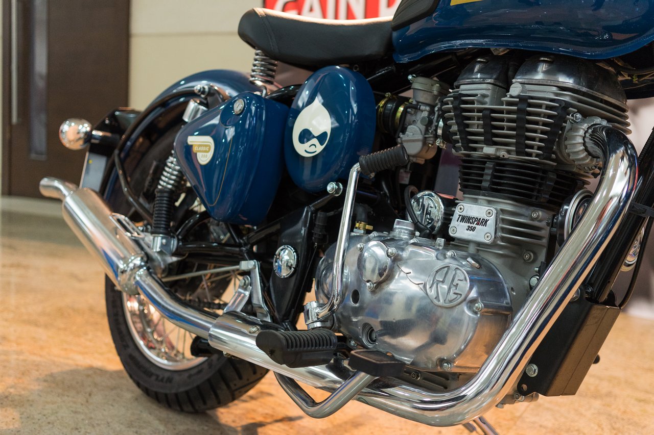 Close-up of a blue Royal Enfield Classic 350 motorcycle, showing its engine, exhaust, and side panel with the Drupal logo engraved.