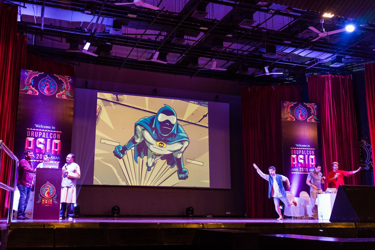 A group of people on stage present a Drupal-themed superhero slide, with some striking playful superhero poses.