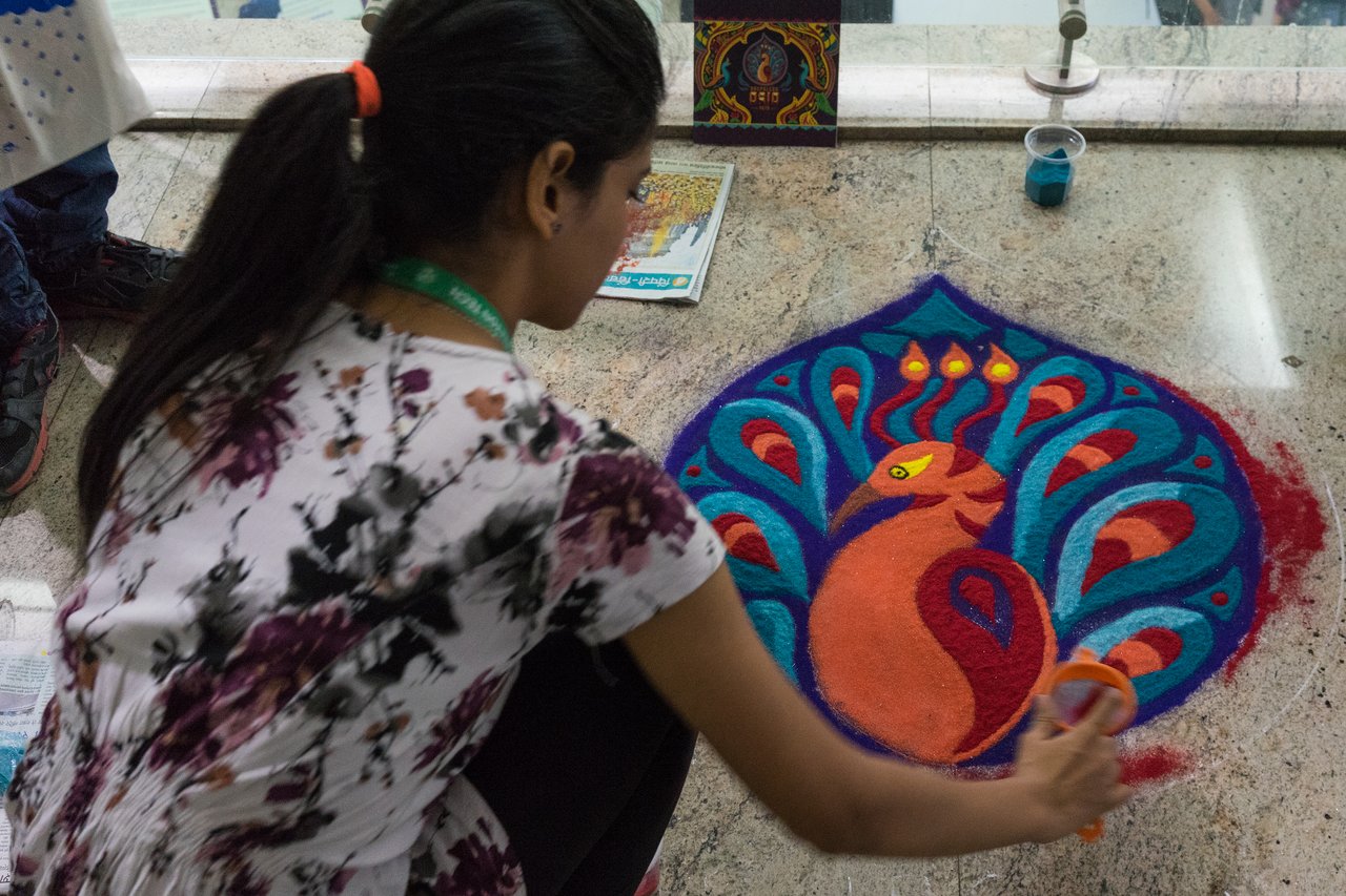 A woman creates a vibrant rangoli design in the shape of Druplicon, the drop-shaped Drupal logo, using colored sand on a marble floor.