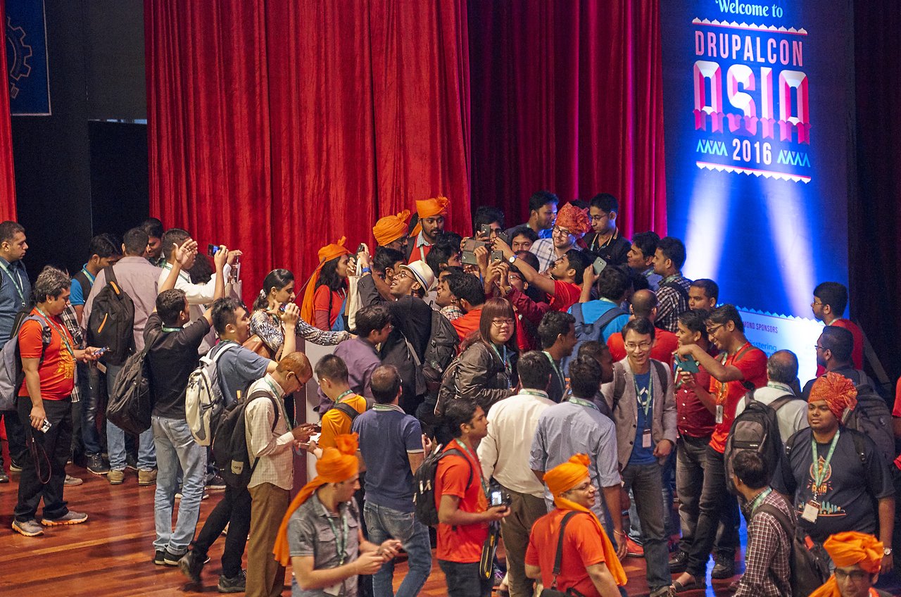 A large group of people at DrupalCon Asia 2016 gather on stage, trying to take a selfie with Dries Buytaert, who remains at the podium where he delivered his talk.