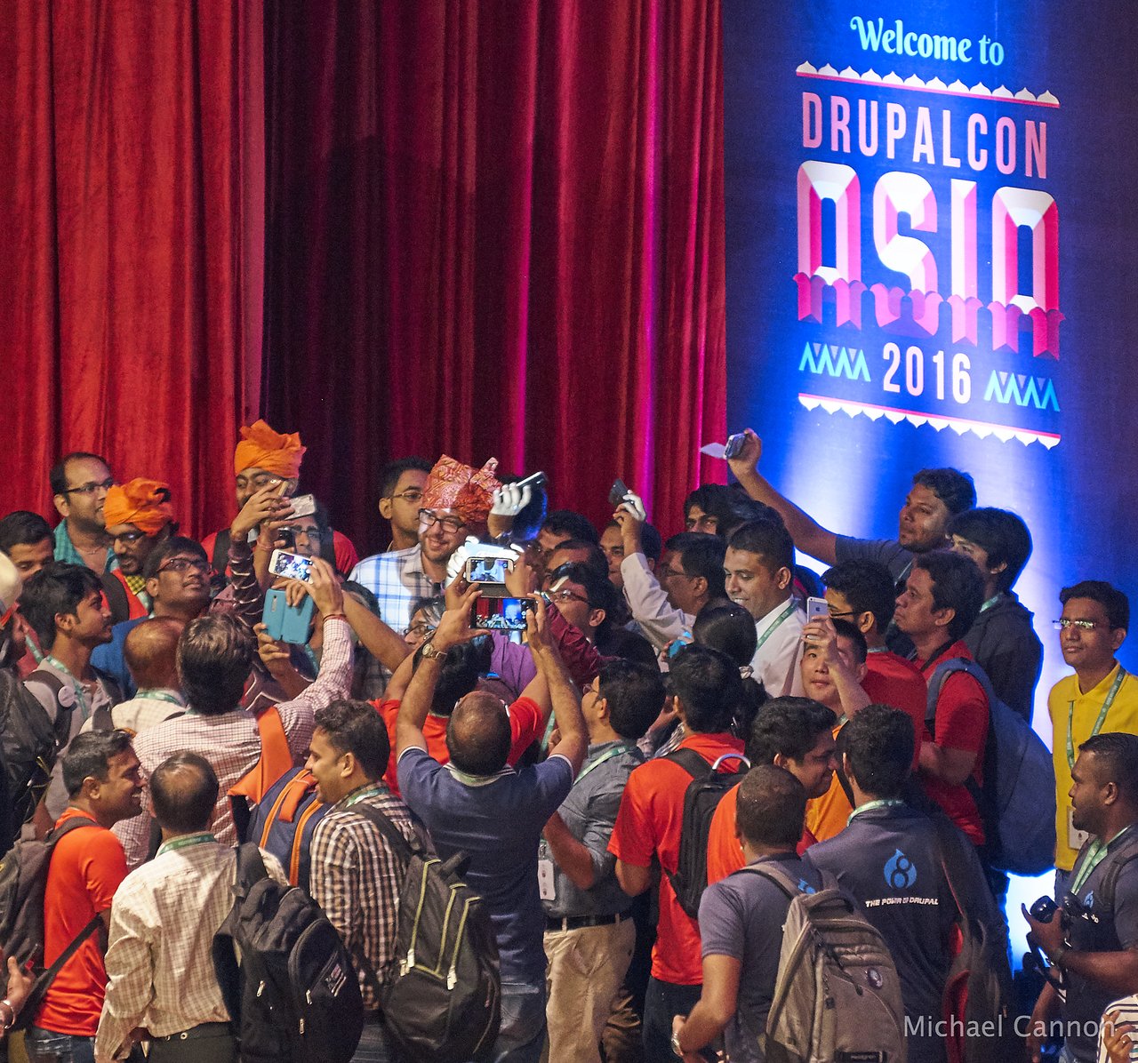 A large group of people at DrupalCon Asia 2016 gather on stage, trying to take a selfie with Dries Buytaert, who remains at the podium where he delivered his talk.
