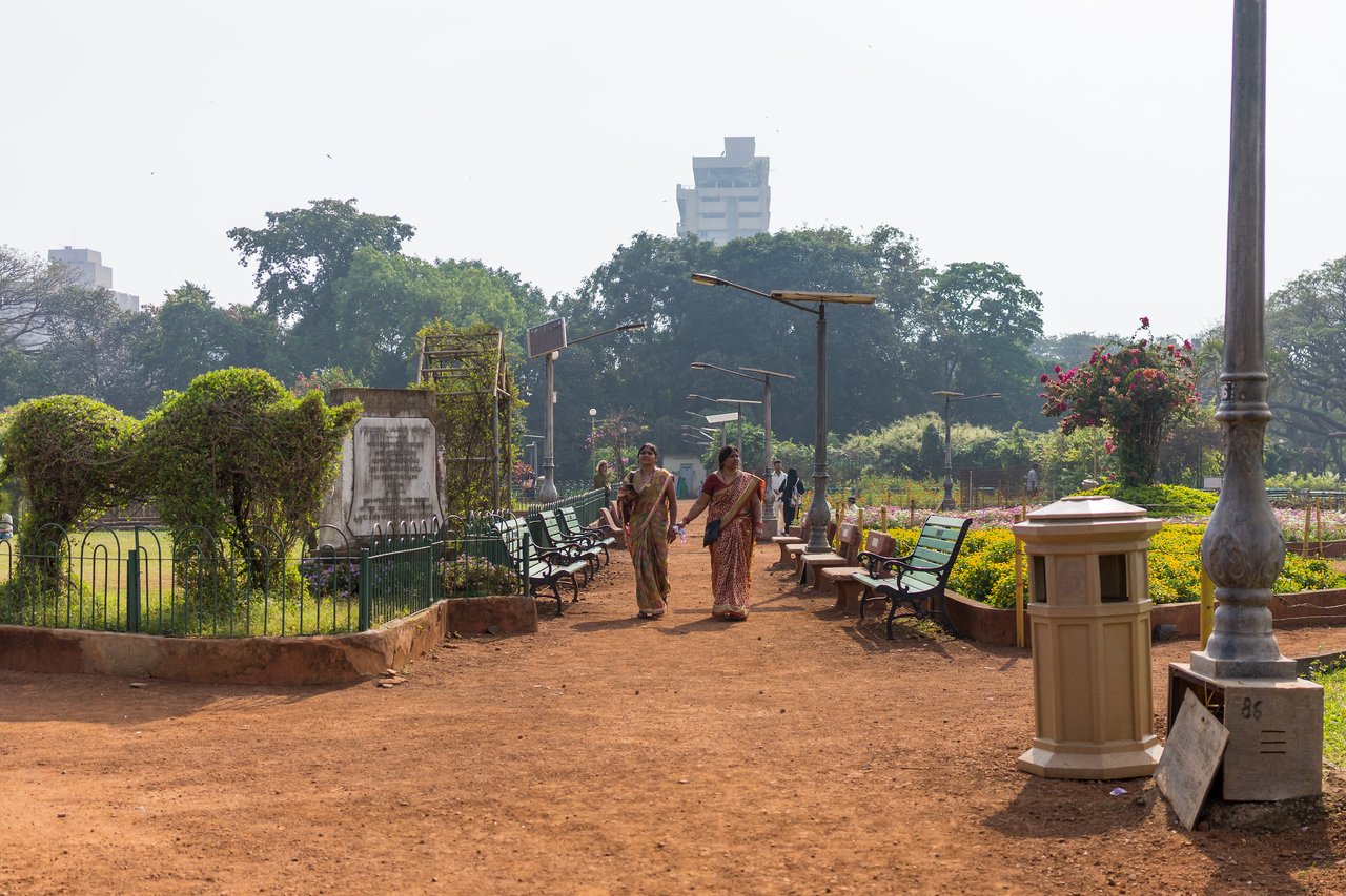 Two women in traditional clothing walk along a dirt path in a park, holding hands and talking.