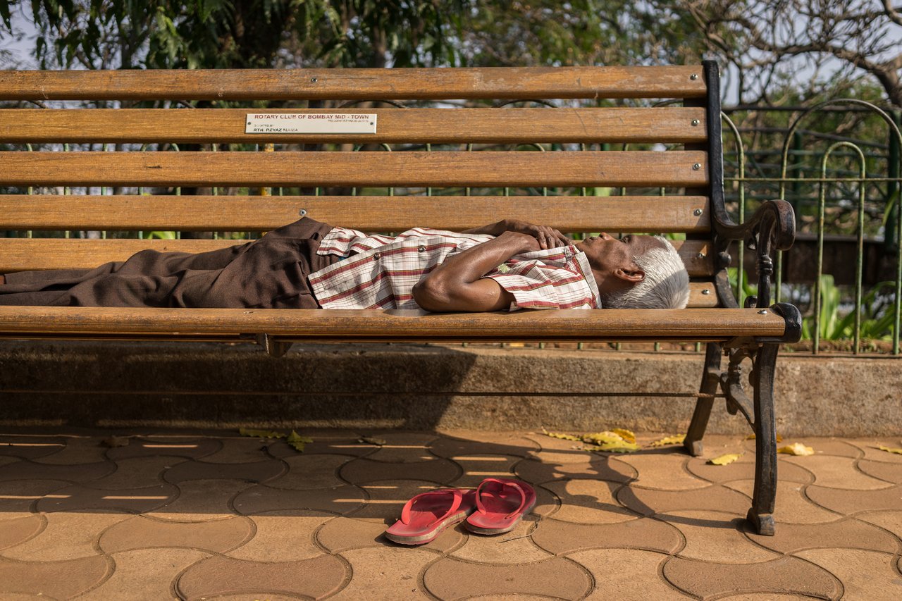 An elderly man lies on a wooden bench with his hands crossed, his red sandals placed on the ground nearby.