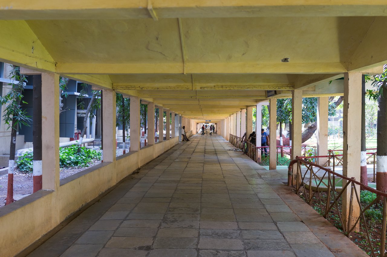 A long, covered walkway at IIT Bombay stretches into the distance, with a few people walking, cycling, and standing by the railing on the right side.