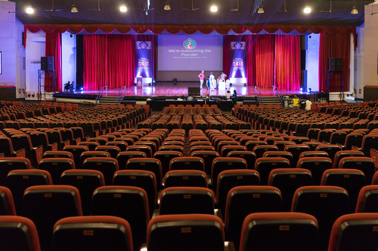An empty auditorium with rows of red seats faces a stage where a small group of people is preparing.