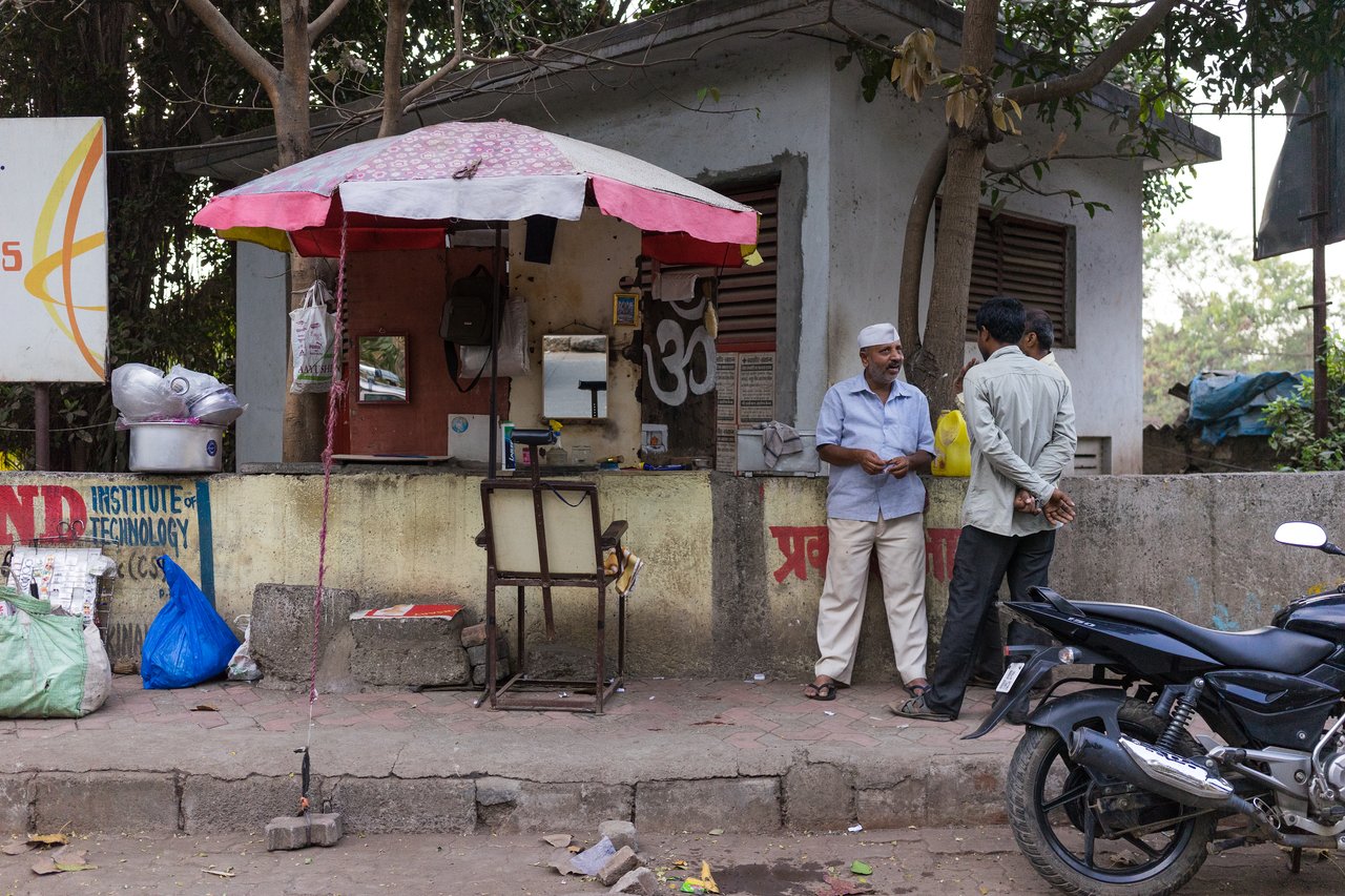 A street barber shop with a chair under an umbrella, while three men stand nearby talking.