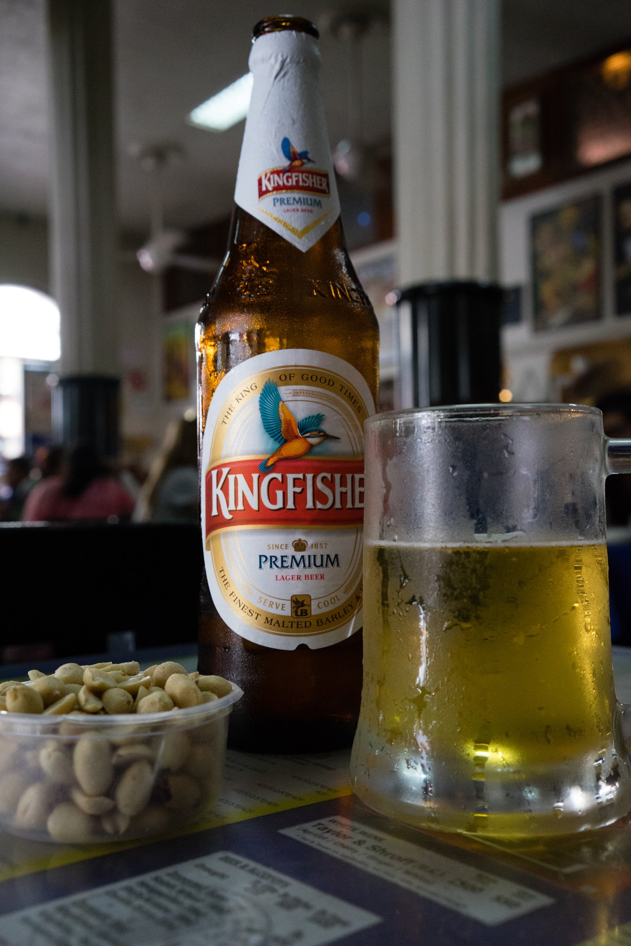 A bottle of Kingfisher beer, a frosty glass of beer, and a small container of peanuts on a table.