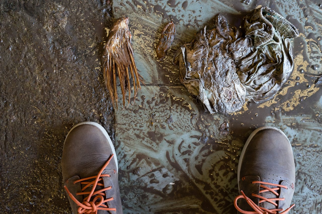 A person wearing brown shoes stands on a muddy floor near a fish head, dirty rag, and debris.