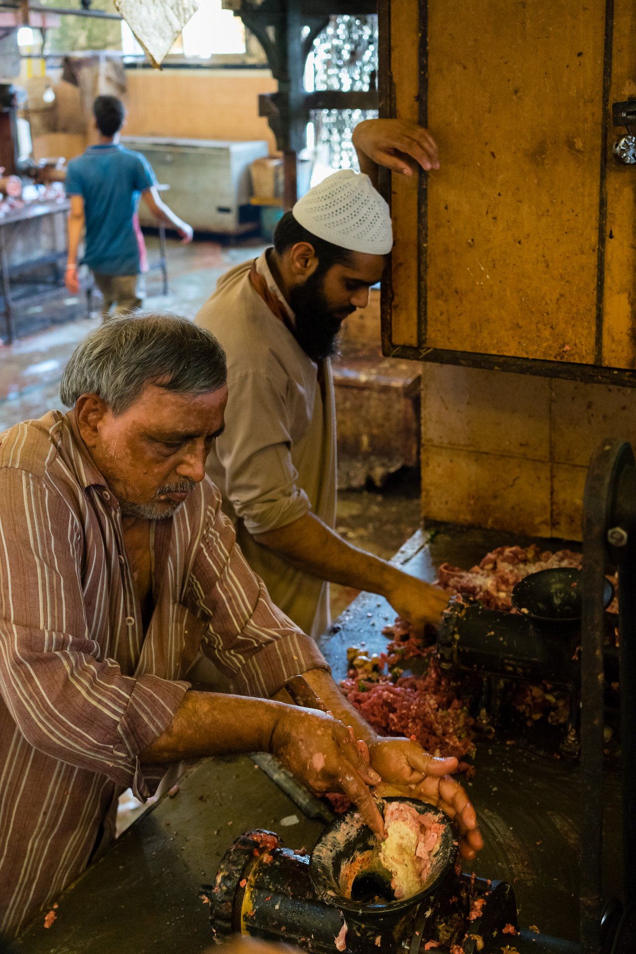 Two men process raw meat using a manual grinder at a butcher shop, pressing it through the machine.