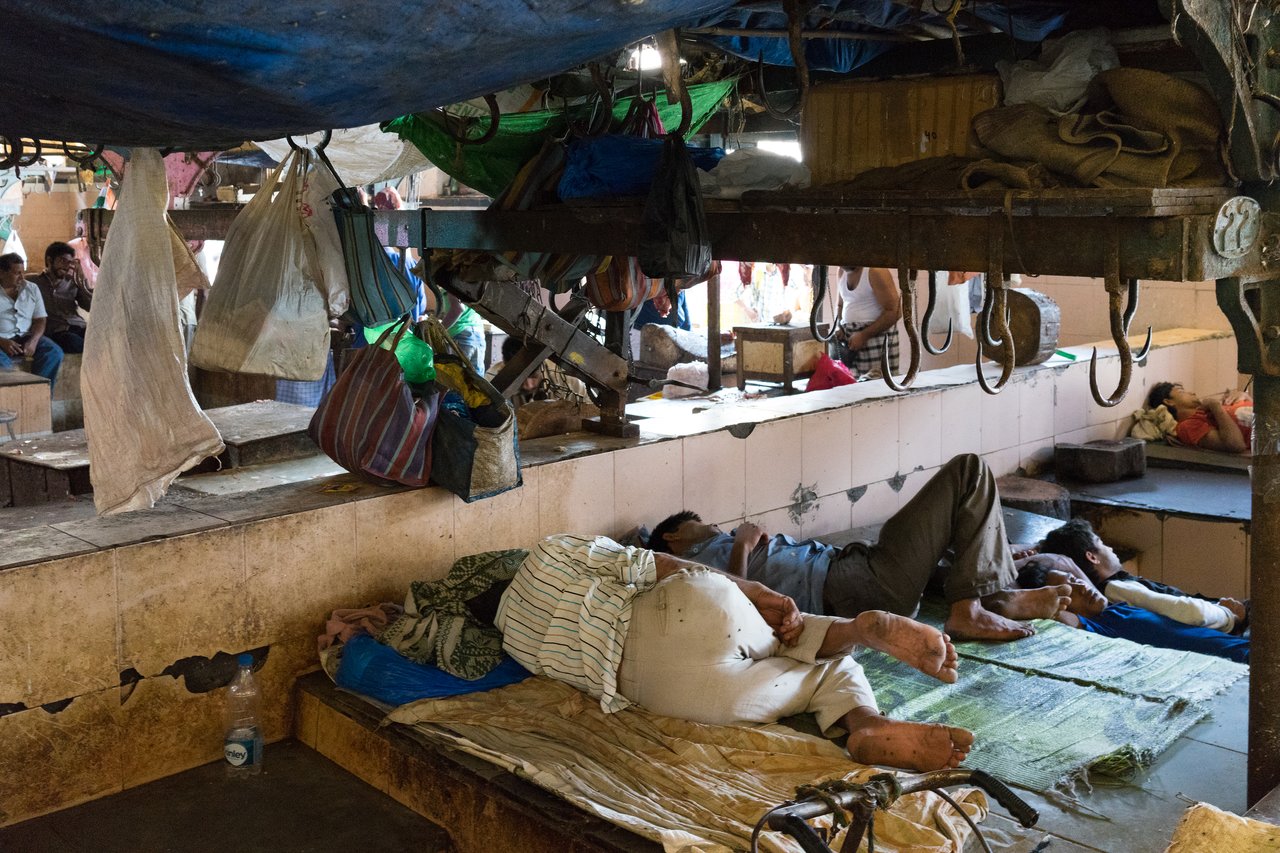 Several people are lying on makeshift beds in a market area, resting among hanging bags and metal hooks.