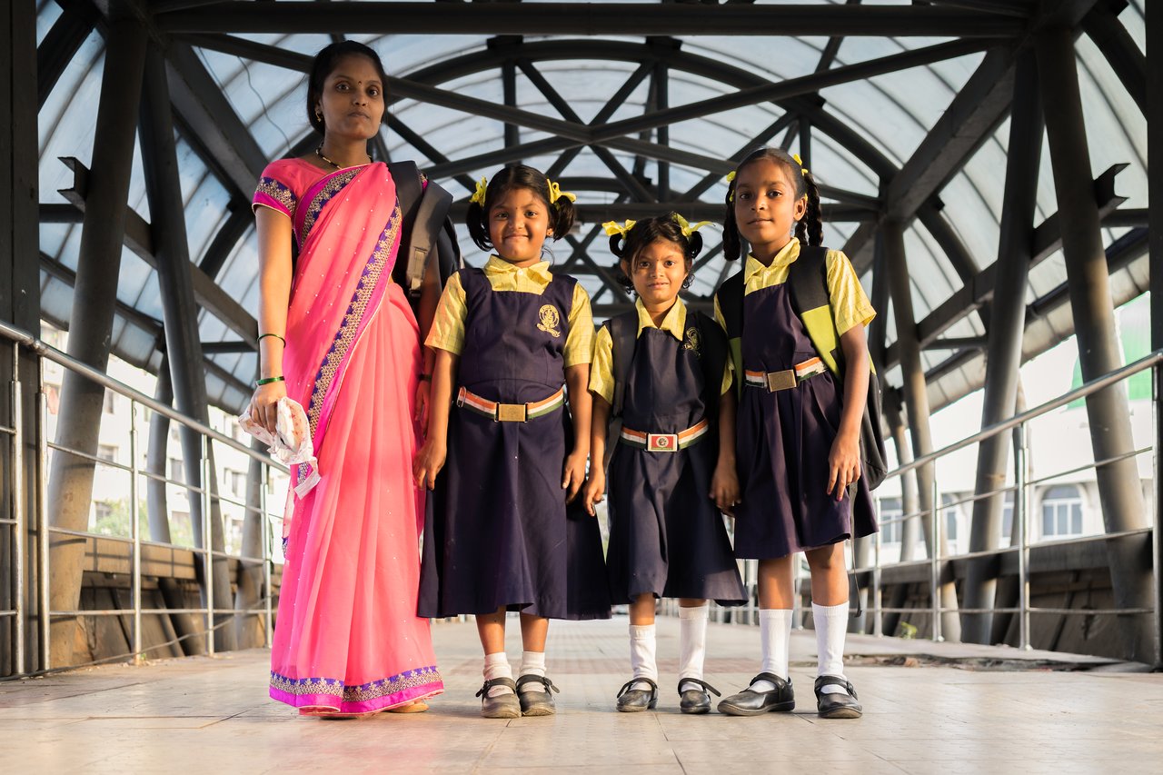 A woman stands with three young schoolgirls in uniforms, all carrying backpacks and looking at the camera.