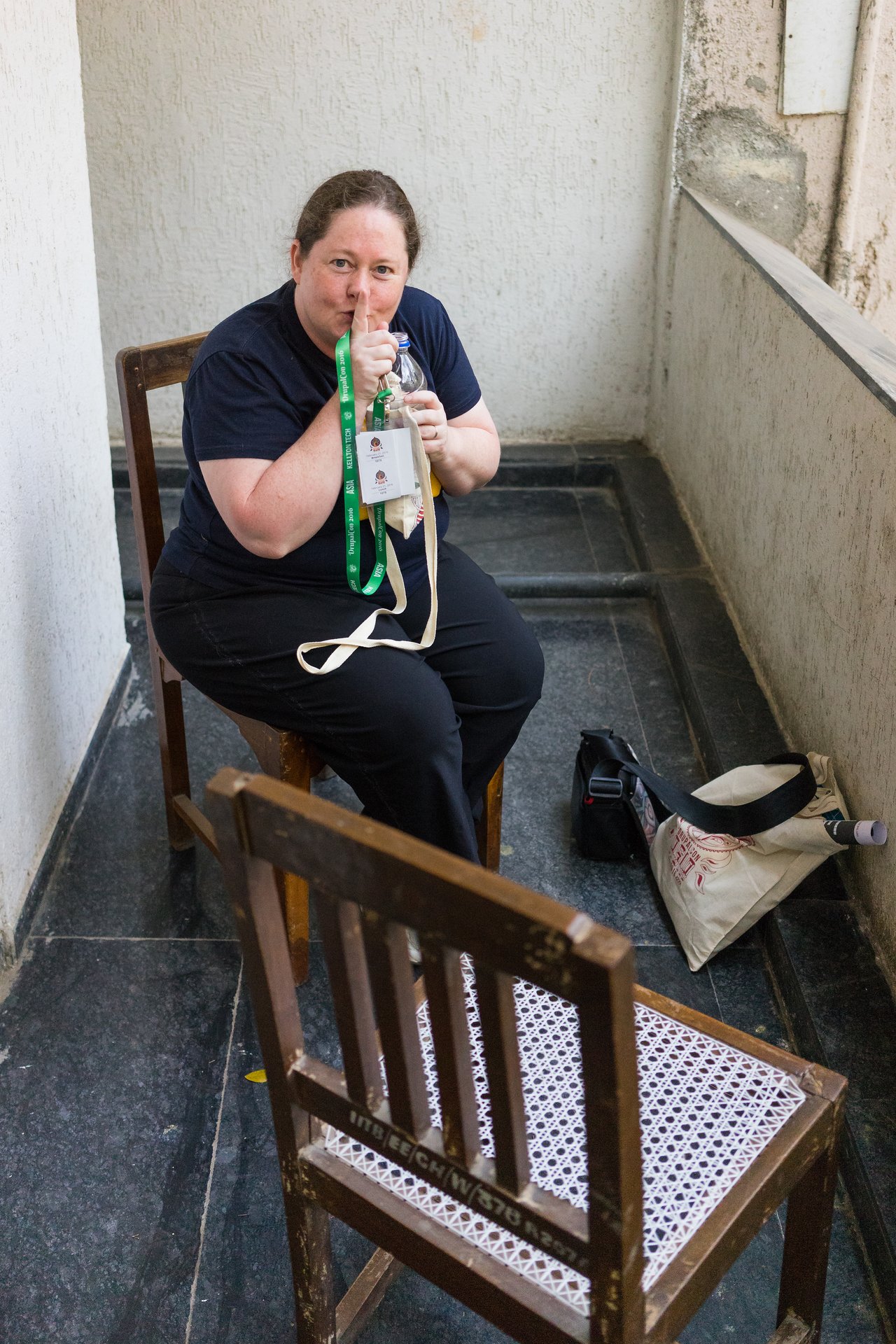 A woman sits on a wooden chair gesturing "shh" with her finger.