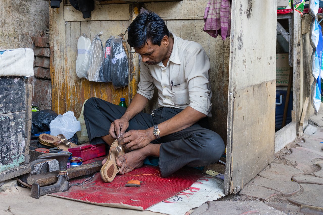A shoemaker sits on the floor, carefully stitching a sandal with a needle and thread.