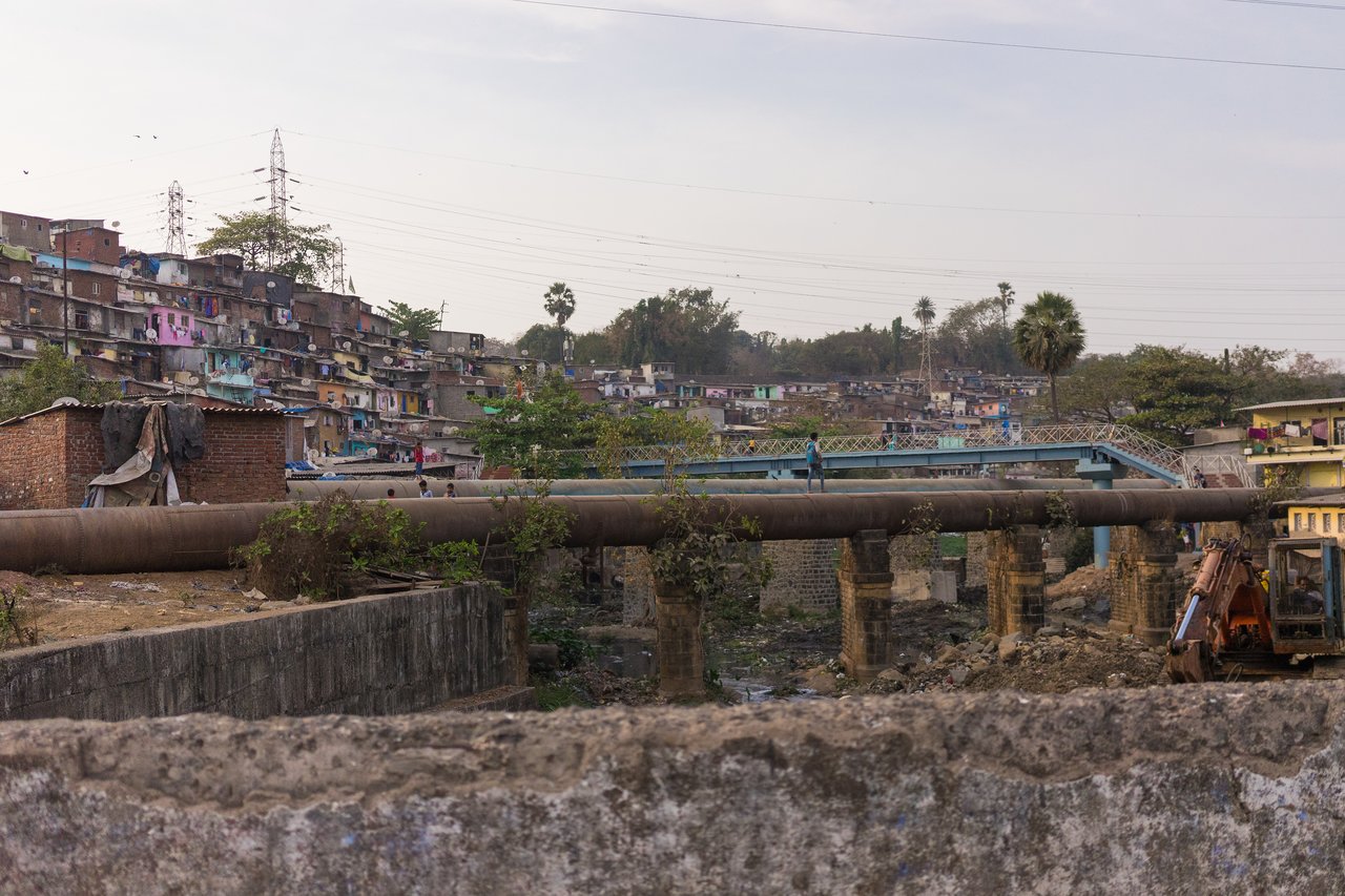 A person walks on a large pipeline over a polluted waterway, with houses and a bridge in the background.