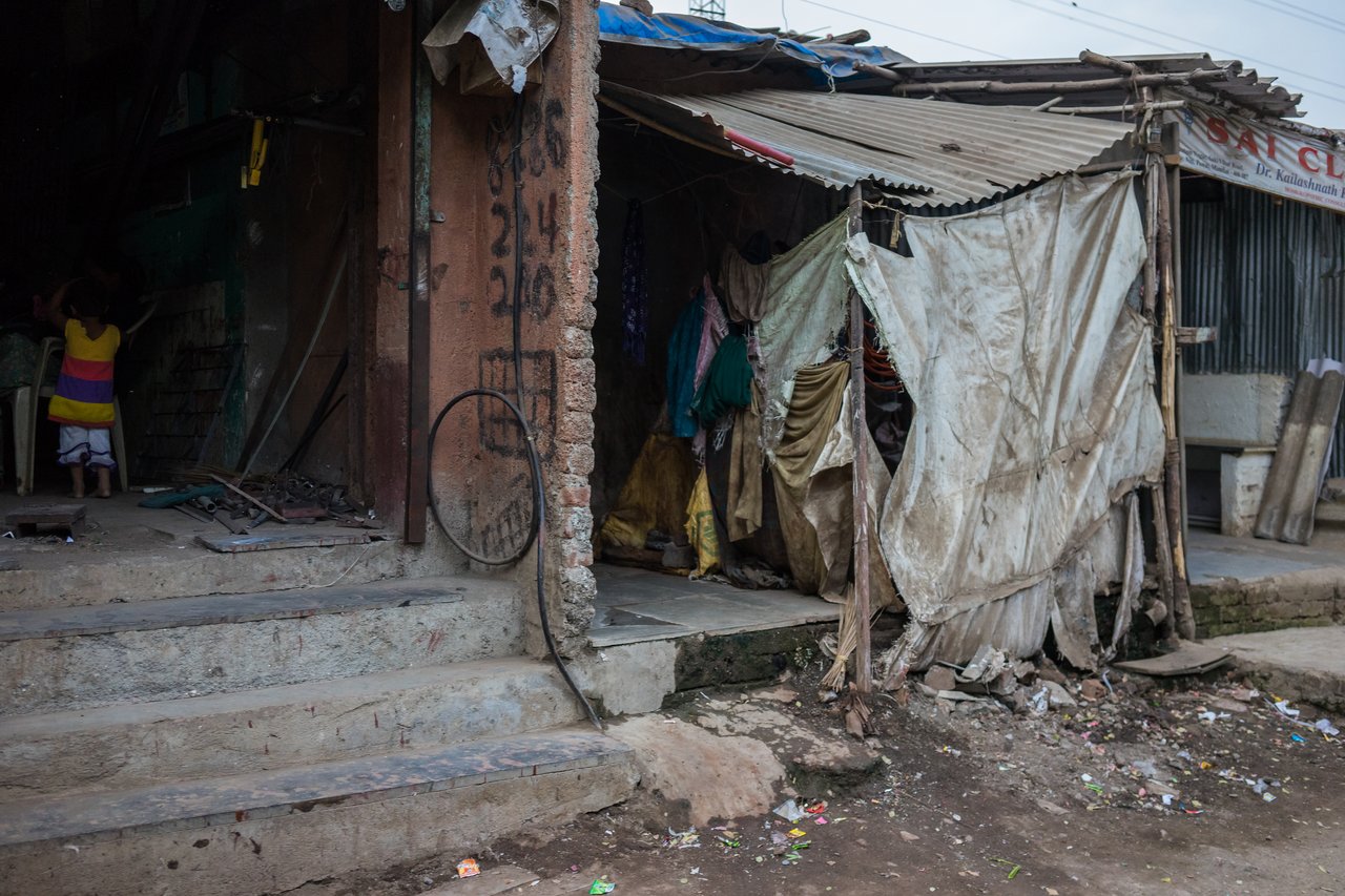 A crumbling slum with makeshift homes and scattered debris with a young child in the shadows.