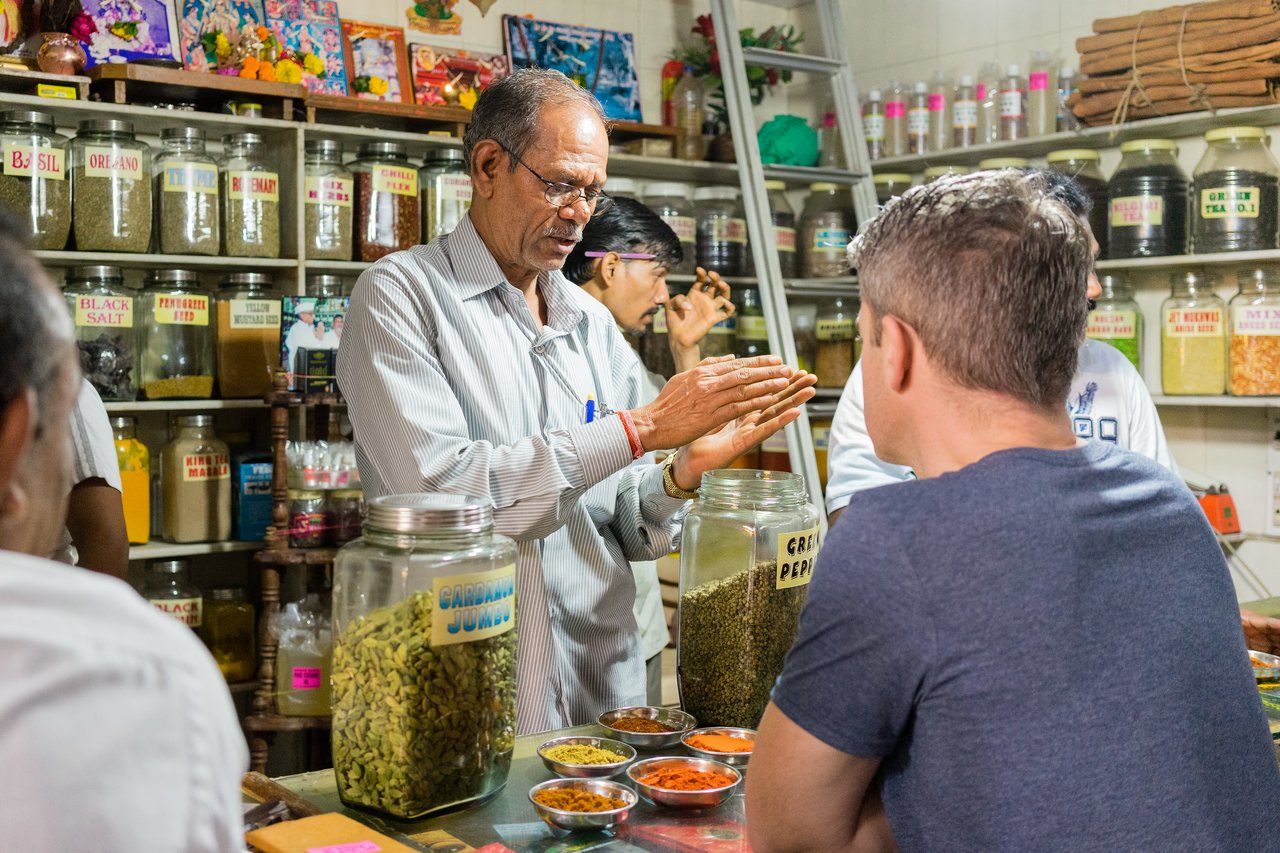 A shopkeeper explains spices to a customer, gesturing with his hands.