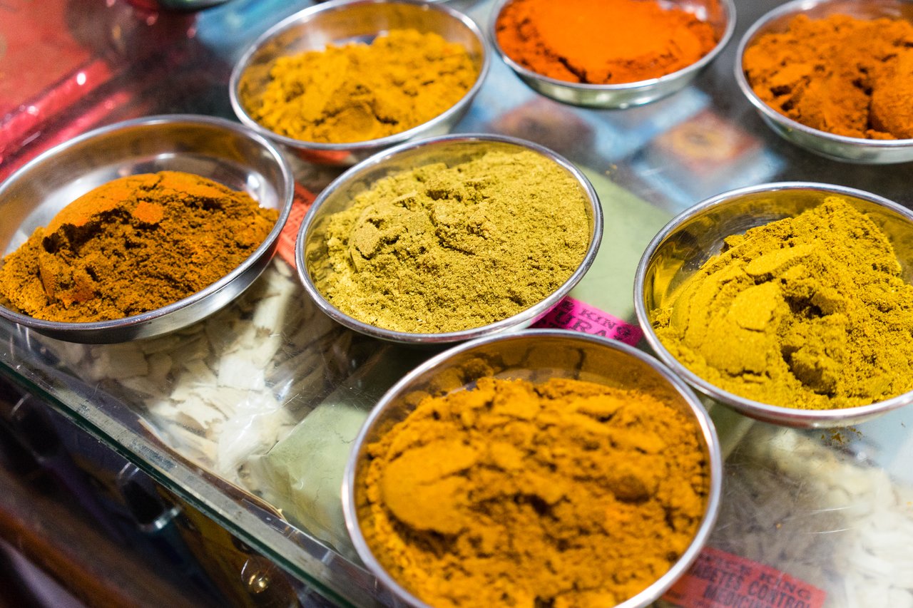 Small metal bowls filled with colorful ground spices are arranged on a glass surface.