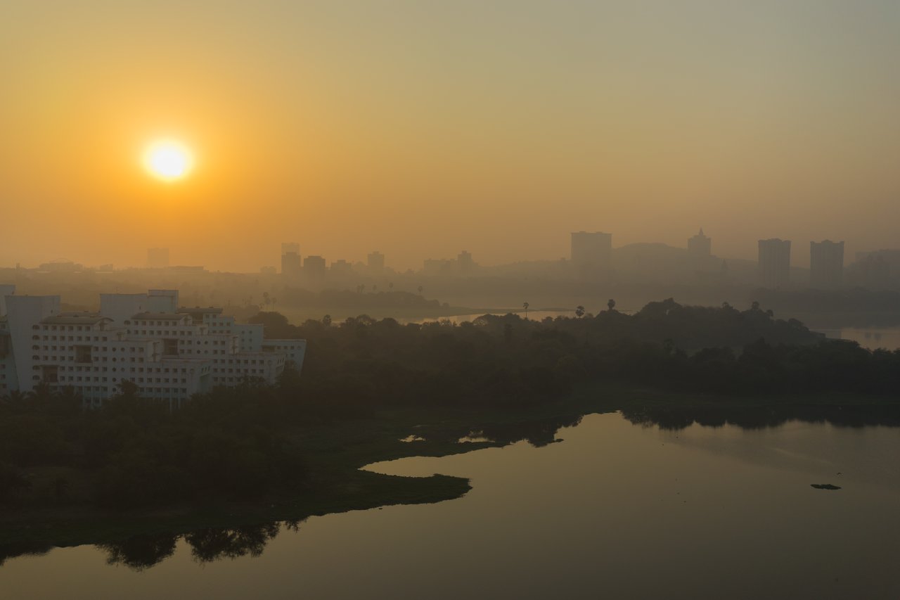 The sun rises over a city skyline, casting a warm, hazy glow on buildings, trees, and a calm lake, intensified by smog or pollution.