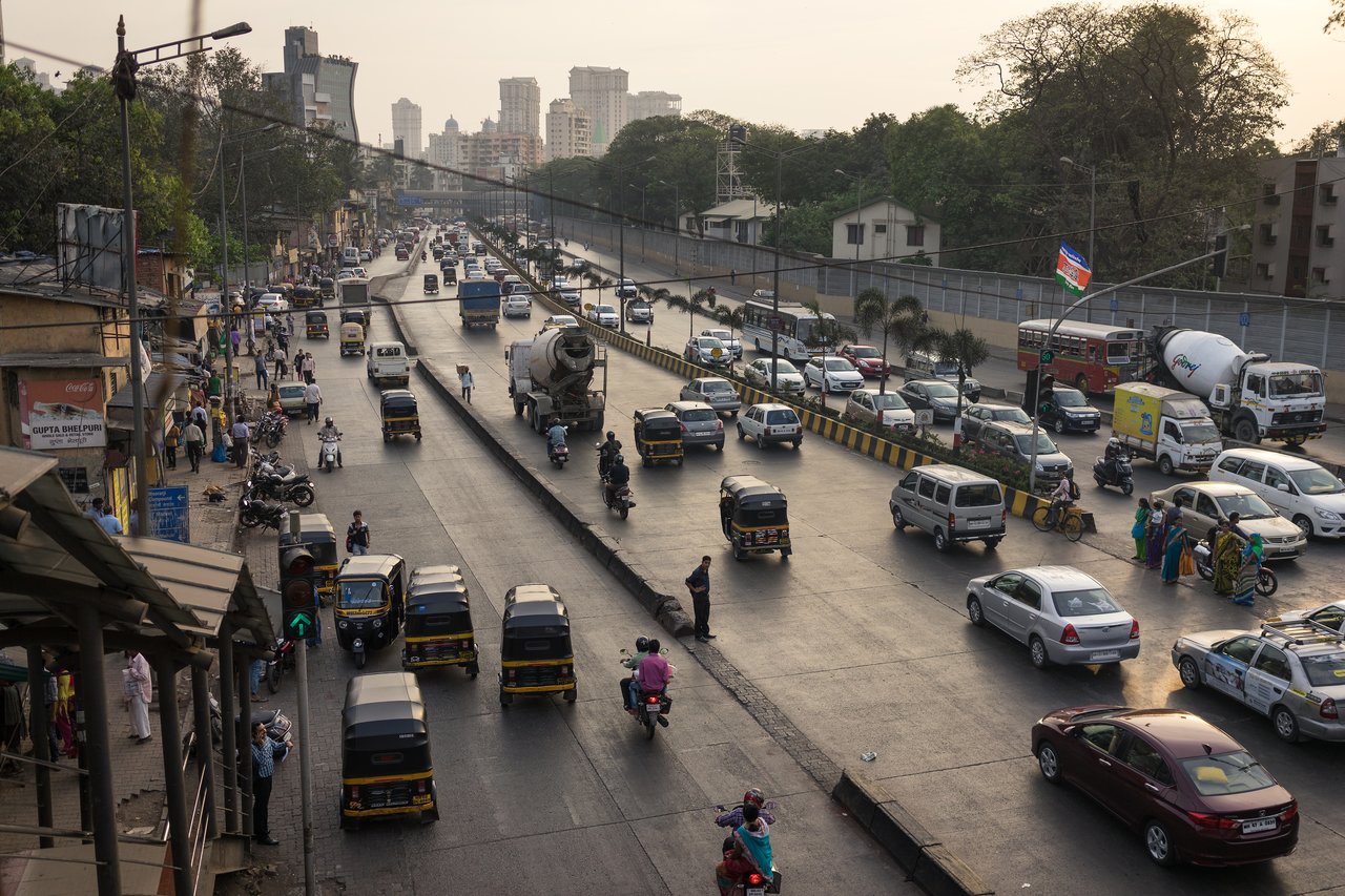 A busy city street with cars, motorcycles, and auto-rickshaws moving in traffic, while pedestrians walk along the sidewalk.