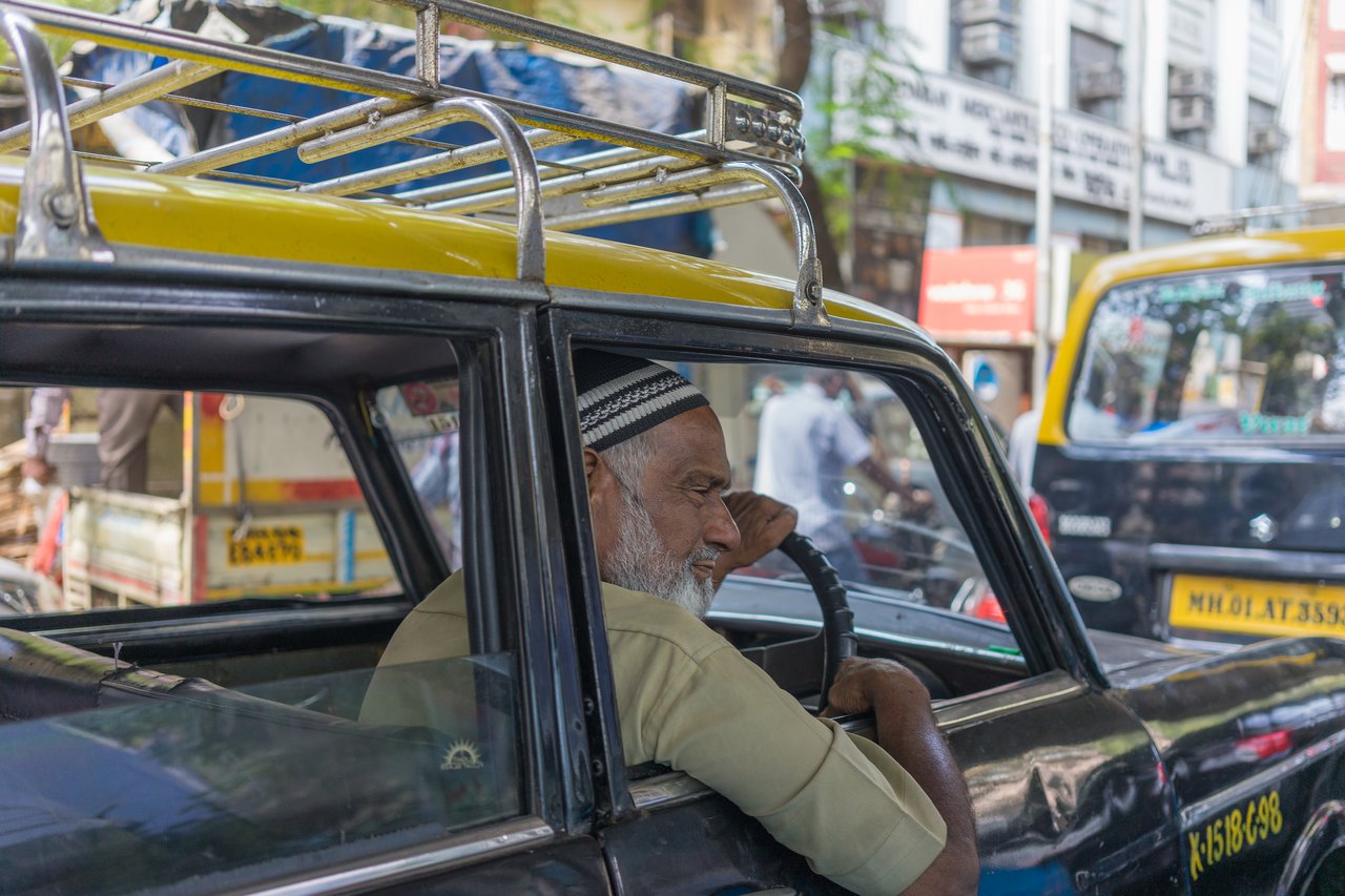 A taxi driver with a gray beard sits in his car, resting his arm on the door while stuck in traffic.
