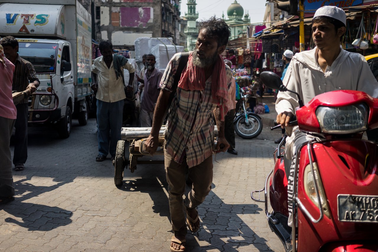 A man pulls a loaded cart through a crowded street while another person rides a red scooter nearby.