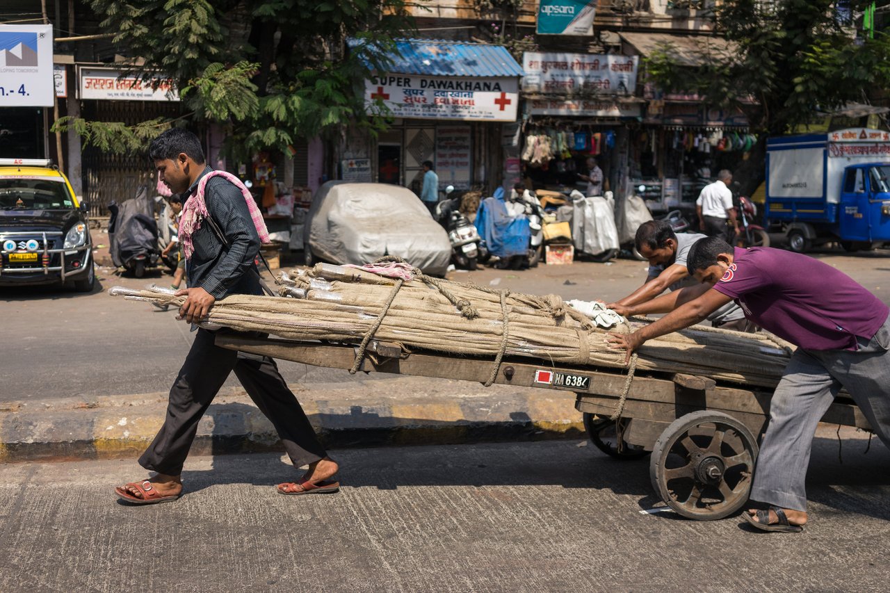 Three men push and pull a heavy wooden cart loaded with bundled sticks on a busy street.