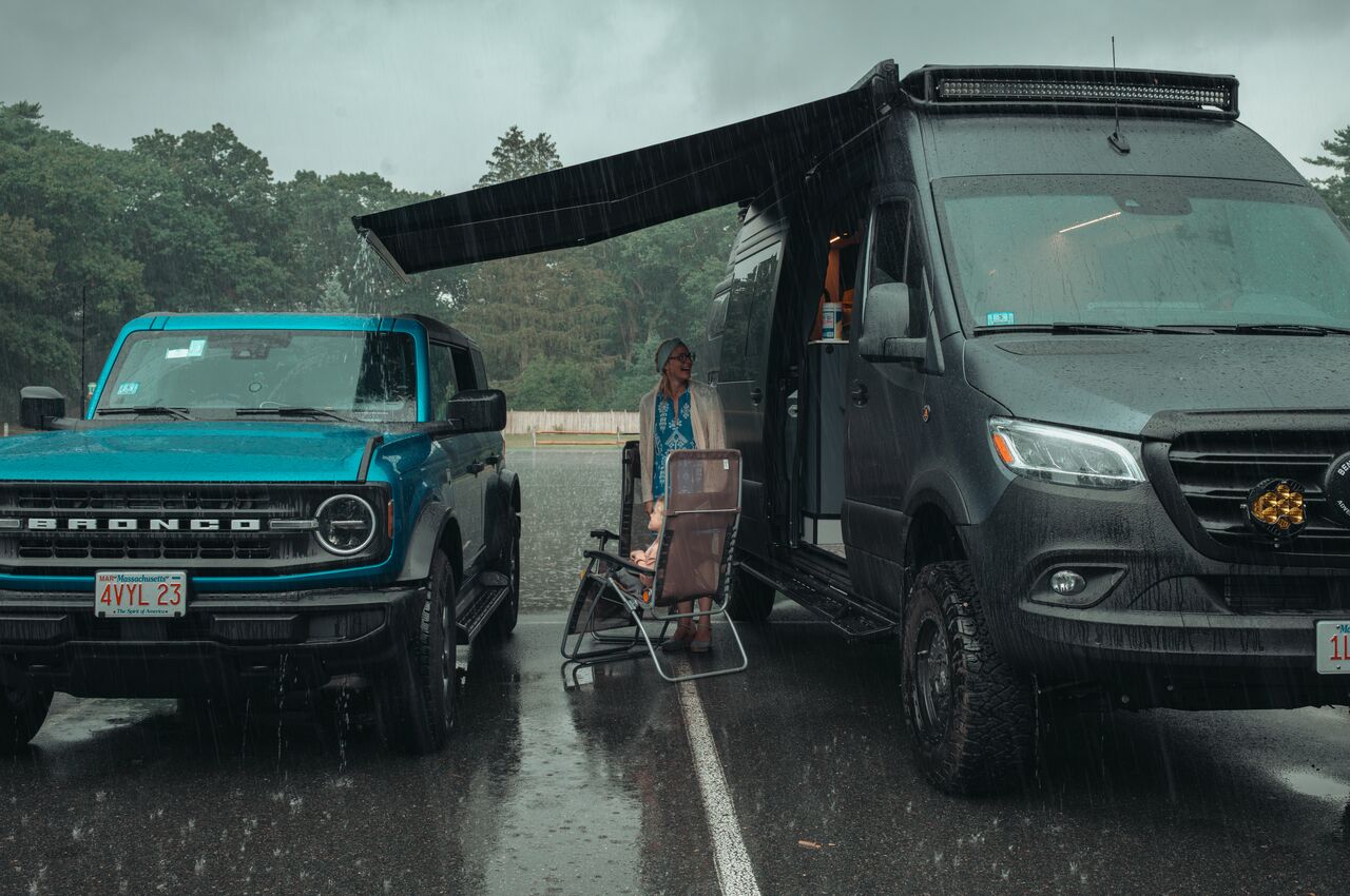 A woman and child sit under an awning between a camper van and SUV, staying dry during heavy rain.