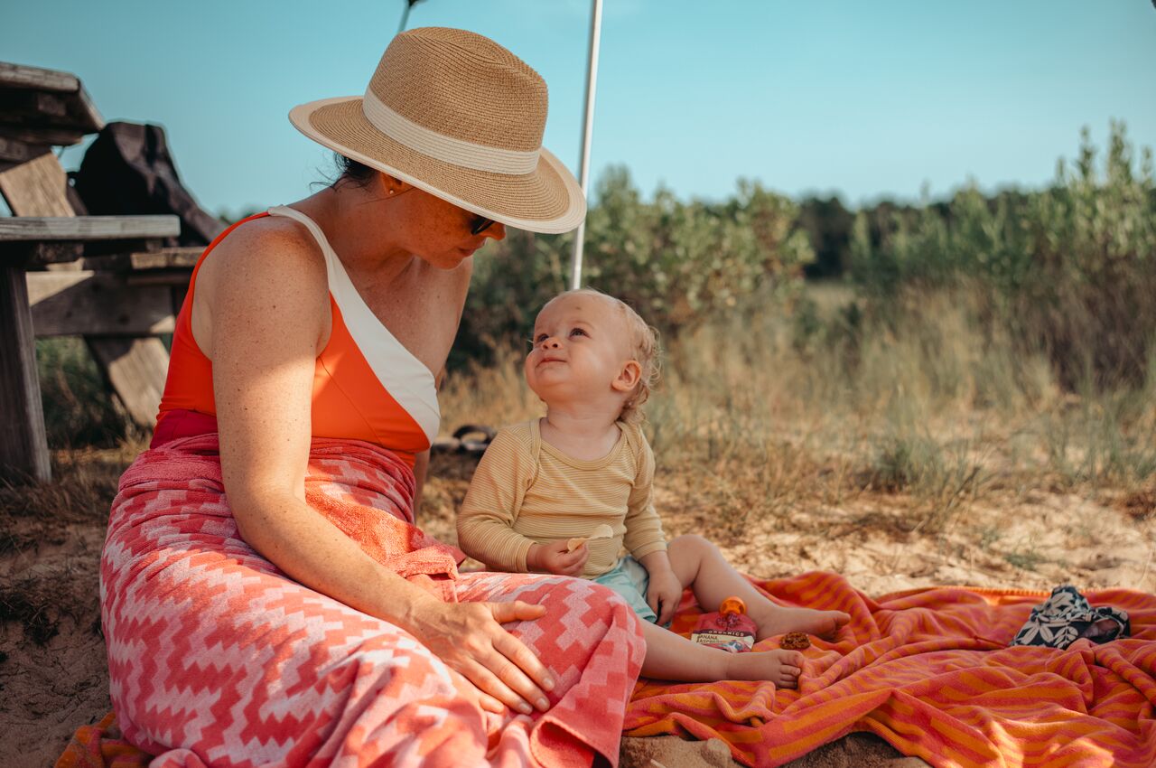 A woman in a sunhat sits beside a toddler on a beach blanket, smiling and looking at each other while sitting in the sand.