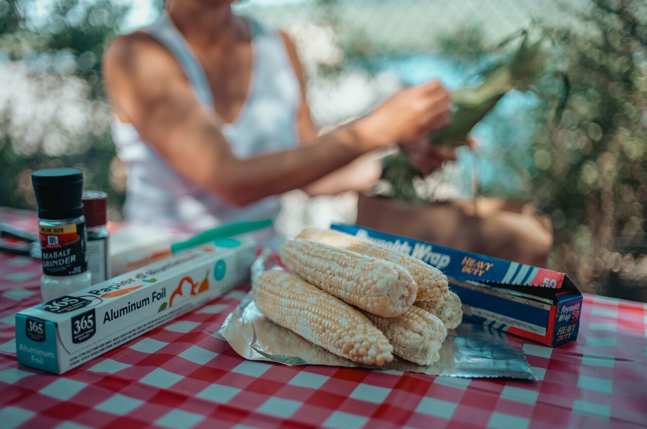 Fresh ears of uncooked corn rest on aluminum foil, while someone in the background husks more corn at a picnic table.