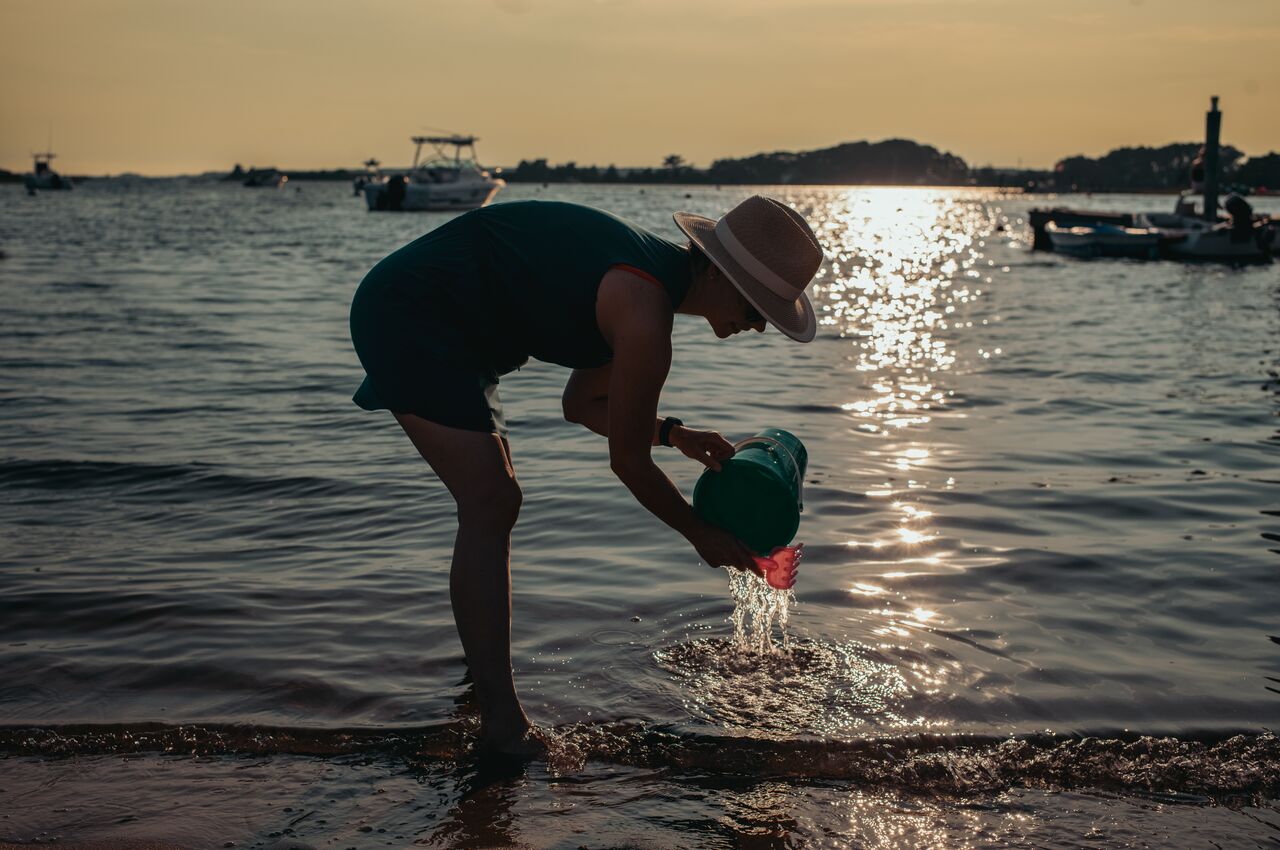 Person wearing a sunhat pours water from a green bucket into the shallow edge of the sea during sunset.