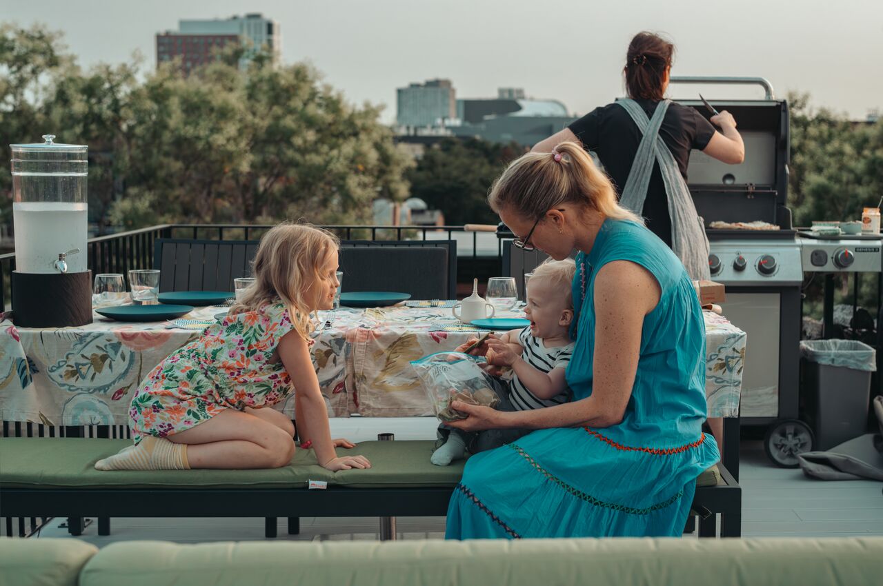A woman sits with two young children at a rooftop table while another person grills food in the background.